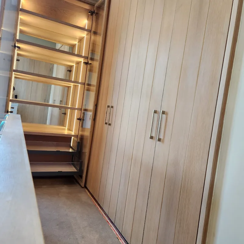 A view of a walk-in closet in natural timber detail with a mirror backed cabinet featruing LED lighting and glass doors for timeless appeal